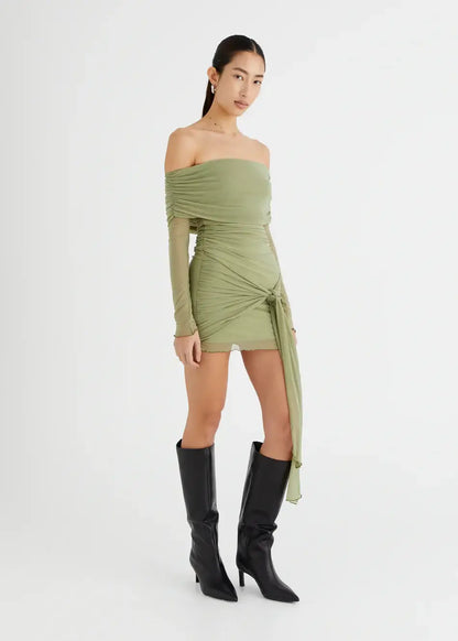 Model wearing Yasmin Off Shoulder Mini Dress in olive green, with sleek black boots and a sarong style skirt for a chic look.