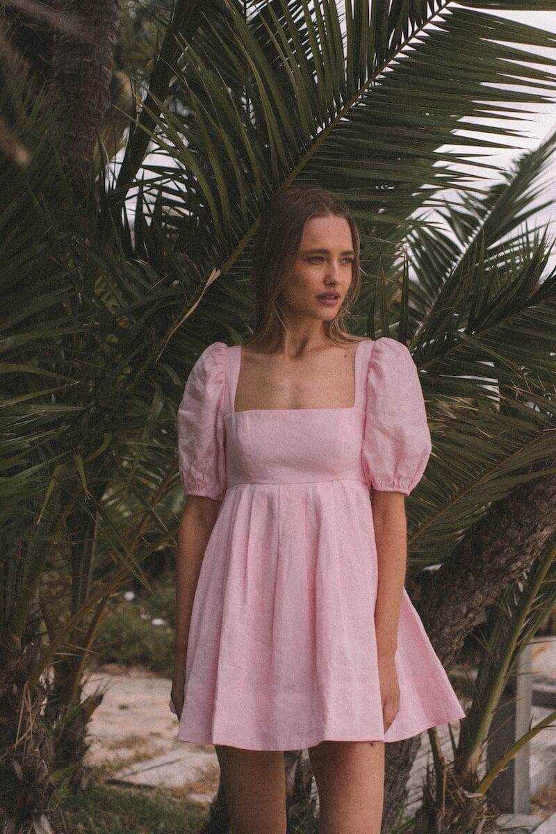 Aerin Open Back Tie Mini Dress in Baby Pink featuring puff sleeves and a pleated skirt against palm trees.