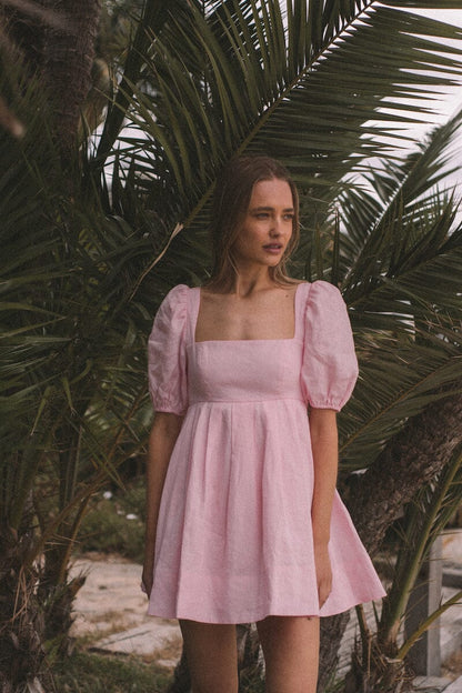 Aerin Open Back Tie Mini Dress in Baby Pink featuring puff sleeves and a pleated skirt against palm trees.